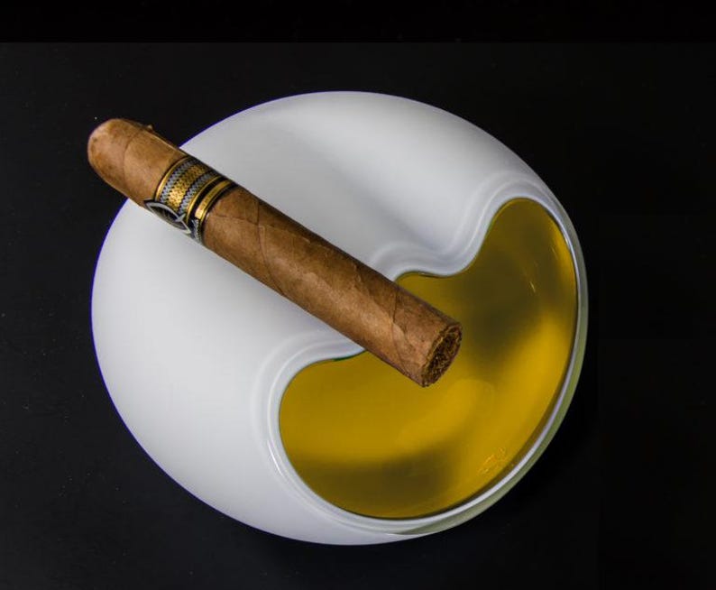 May include: White ceramic ashtray with a yellow, liquid-filled compartment. A brown cigar rests on the edge of the ashtray.