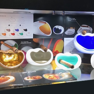May include: A display of various ceramic and glass ashtrays in different colors and shapes. The ashtrays are designed to hold cigars and feature a unique, modern aesthetic. The display also includes a round tray with the word "Cevitrum" printed on it.