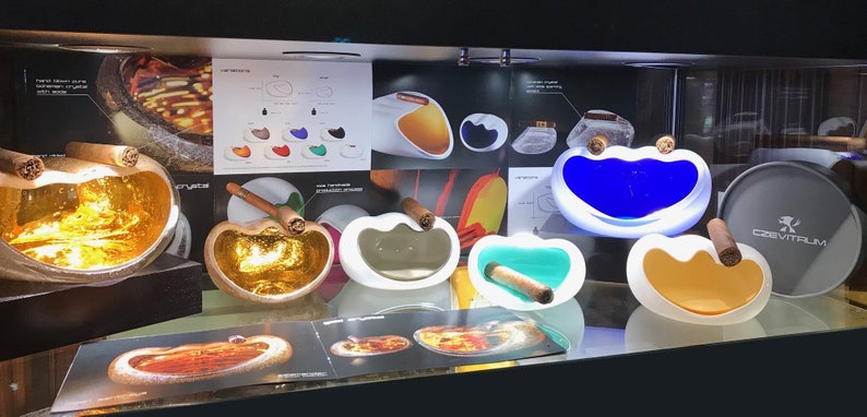 May include: A display of various ceramic and glass ashtrays in different colors and shapes. The ashtrays are designed to hold cigars and feature a unique, modern aesthetic. The display also includes a round tray with the word "Cevitrum" printed on it.