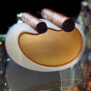 May include: A white ceramic cigar holder with two brown cigars resting in the grooves. The holder has a smooth, rounded shape and a gold-colored interior.