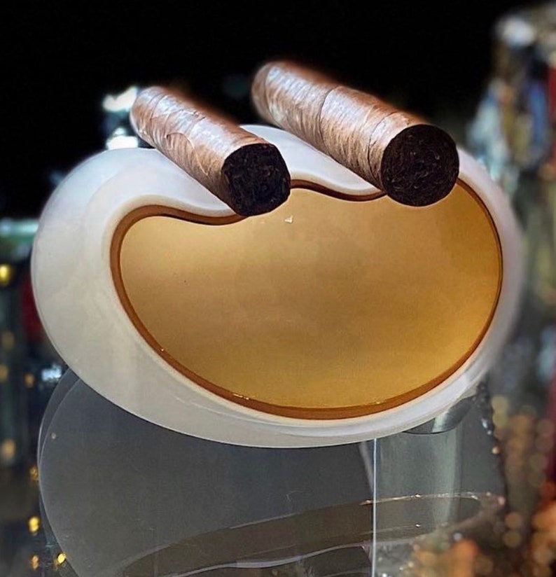 May include: A white ceramic cigar holder with two brown cigars resting in the grooves. The holder has a smooth, rounded shape and a gold-colored interior.