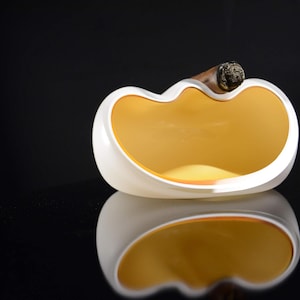 May include: White ceramic ashtray with a yellow interior and a single cigar resting on the rim.