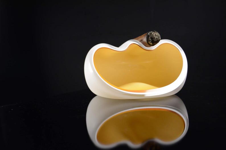 May include: White ceramic ashtray with a yellow interior and a single cigar resting on the rim.