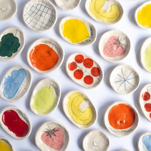 Könnte beinhalten: Eine Sammlung kleiner, ovaler Keramikschalen mit handgemalten Illustrationen verschiedener Obst- und Gemüsesorten sowie einfachen, strichgezeichneten Smileys. Die Schalen sind in leuchtenden Farben wie Rot, Gelb, Orange, Grün und Blau glasiert.