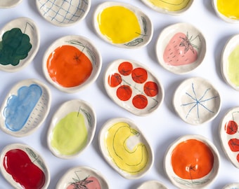 May include: A collection of small, oval-shaped ceramic dishes featuring hand-painted illustrations of various fruits, vegetables, and simple line-drawn smiley faces. The dishes are glazed in vibrant colors including red, yellow, orange, green, and blue.