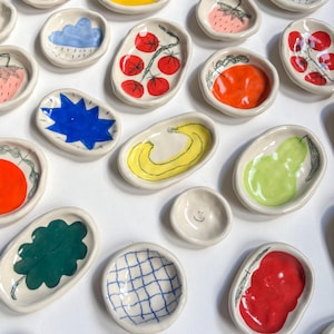 Könnte beinhalten: Eine Sammlung kleiner, handbemalter Keramikschalen. Jede Schale hat ein einzigartiges Design, darunter Früchte, Gemüse und abstrakte Muster in leuchtenden Farben wie Rot, Gelb, Grün und Blau. Die Schalen variieren in Form und Größe.