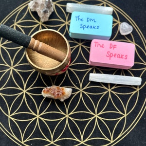 May include: A black mat with a gold flower of life design. A singing bowl with a wooden striker, two white selenite sticks, two pink and blue sticky notes with the text "The DM Speaks" and "The DF Speaks", and two clear quartz crystals are arranged on the mat.