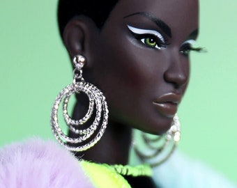Può includere: Una bambola di moda con capelli corti neri, che indossa una giacca di pelliccia sintetica viola, un top patchwork verde neon e bianco e pantaloni neri. La bambola ha la pelle scura, eyeliner nero e grandi orecchini a cerchio argentati.