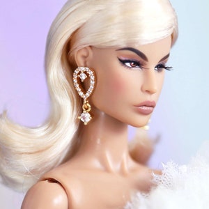 Op de afbeelding: Een blonde pop met een witte veren- en kantjurk en gouden oorbellen met heldere stenen. De pop kijkt naar rechts.