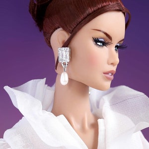 May include: A doll with long, dark brown hair styled in a bun, wearing a white, ruffled blouse. The doll has a pearl earring with a crystal embellishment.