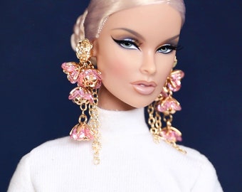 May include: A doll with blonde hair and a white turtleneck sweater. The doll is wearing large pink and gold earrings.