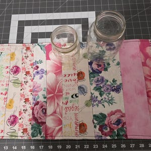 May include: A rectangular fabric placemat featuring vertical strips of various patterns including pink flowers, musical instruments, and text, with two clear glass jars of different sizes placed on top of the quilted surface.