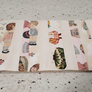 May include: A rectangular fabric placemat featuring a vintage paper doll theme with illustrations of children, various outfits, hats, and floral accents printed in soft pastel colors on a cream background with vertical panel stitching throughout.