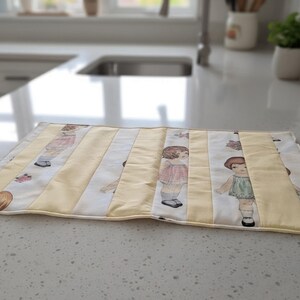 May include: A rectangular fabric placemat featuring alternating vertical panels of solid pale yellow fabric and white fabric printed with illustrations of children in vintage-style clothing. The placemat has a stitched border and a quilted texture.