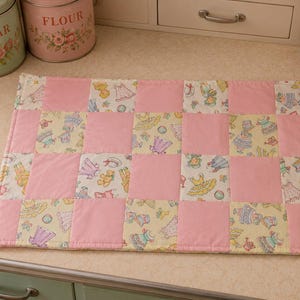 May include: A rectangular quilted placemat featuring a checkerboard pattern of solid light pink fabric squares alternating with squares of cream-colored fabric printed with small illustrations of dresses, hats, and teddy bears in pastel colors.