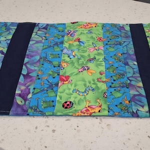 May include: A rectangular fabric placemat featuring a pieced design with alternating vertical strips. The pattern includes bright green sections with colorful insects and blue sections with various creatures, separated by solid dark navy blue fabric bands.