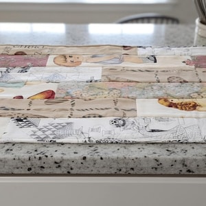 May include: A rectangular fabric placemat featuring a patchwork design of various printed panels. The patterns include illustrations of dolls, floral motifs, and geometric shapes in a color palette of beige, white, red, yellow, and muted pink tones.