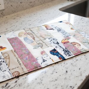 May include: A rectangular fabric mat featuring a patchwork design with various printed illustrations of vintage-style dolls, floral patterns, and decorative motifs in shades of pink, blue, black, white, and tan with stitched quilted borders.