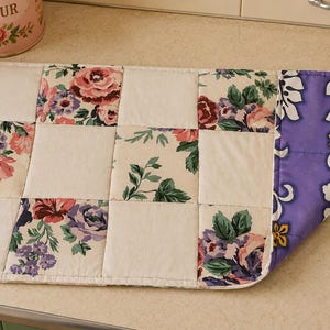 May include: A rectangular quilted placemat featuring a patchwork design of solid cream squares and floral print squares with pink, purple, and green roses. The underside of the placemat is visible, showing a purple fabric with a white hibiscus pattern.