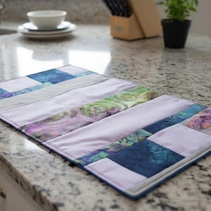 May include: A rectangular quilted placemat featuring a patchwork design of solid light lavender fabric strips alternating with patterned fabric sections in shades of deep blue, teal, purple, and green with swirling and mottled textures.