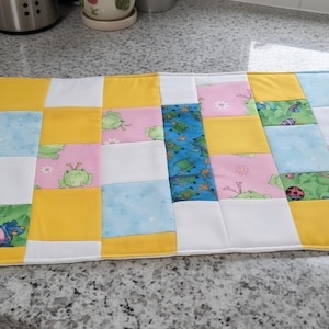 May include: A rectangular quilted placemat featuring a patchwork design of square fabric blocks in yellow, white, light blue, and pink. Some blocks display a green frog pattern, while a central vertical strip features a blue turtle print.
