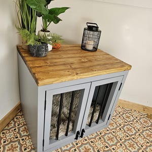 K&ouml;nnte beinhalten: Eine graue Hundebox mit einer Holzplatte und zwei vergitterten T&uuml;ren. Die Box hat ein Bett aus Kunstfell im Inneren. Die Oberseite ist mit Pflanzen und einer schwarzen Metalllaterne dekoriert. Die Box steht auf einem gemusterten Fliesenboden.
