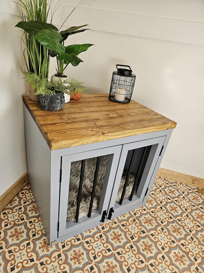 K&ouml;nnte beinhalten: Eine graue Hundebox mit einer Holzplatte und zwei vergitterten T&uuml;ren. Die Box hat ein Bett aus Kunstfell im Inneren. Die Oberseite ist mit Pflanzen und einer schwarzen Metalllaterne dekoriert. Die Box steht auf einem gemusterten Fliesenboden.