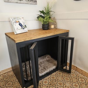 K&ouml;nnte beinhalten: Eine schwarze Hundebox mit einer Holzplatte und zwei T&uuml;ren. Die Box hat vertikale St&auml;be an den T&uuml;ren und ein Kunstfellbett im Inneren. Ein gerahmter Tukan-Druck und Topfpflanzen stehen oben drauf. Ein Elefantenkunstdruck h&auml;ngt an der Wand.