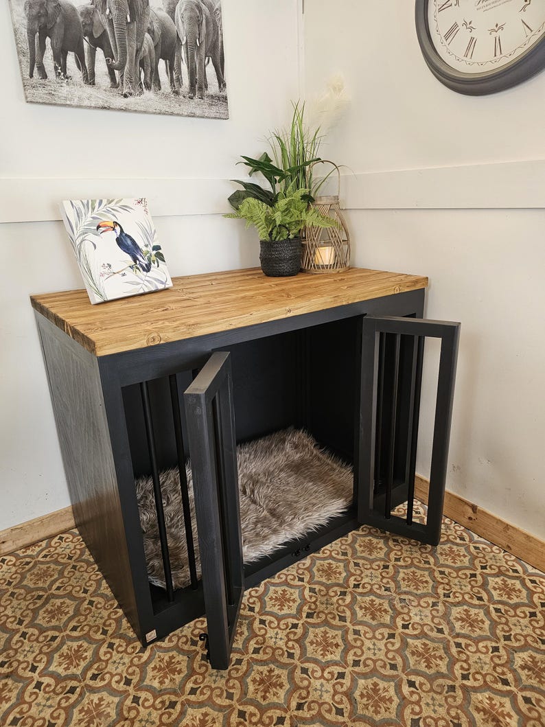 K&ouml;nnte beinhalten: Eine schwarze Hundebox mit einer Holzplatte und zwei T&uuml;ren. Die Box hat vertikale St&auml;be an den T&uuml;ren und ein Kunstfellbett im Inneren. Ein gerahmter Tukan-Druck und Topfpflanzen stehen oben drauf. Ein Elefantenkunstdruck h&auml;ngt an der Wand.