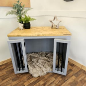 K&ouml;nnte beinhalten: Eine Hundebox aus Holz mit hellgrauem Rahmen und einer Oberseite aus Naturholz. Die Box hat zwei T&uuml;ren mit schwarzen St&auml;ben und ein flauschiges Kunstfellbett im Inneren. Eine Uhr und ein dekorativer Stern befinden sich oben.