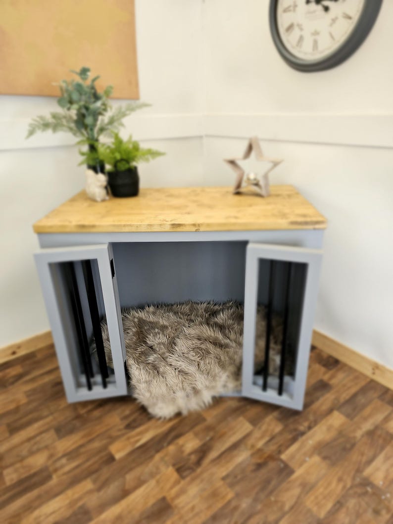 K&ouml;nnte beinhalten: Eine Hundebox aus Holz mit hellgrauem Rahmen und einer Oberseite aus Naturholz. Die Box hat zwei T&uuml;ren mit schwarzen St&auml;ben und ein flauschiges Kunstfellbett im Inneren. Eine Uhr und ein dekorativer Stern befinden sich oben.