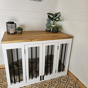 K&ouml;nnte beinhalten: Eine wei&szlig;e Hundebox mit einer Holzplatte und schwarzen St&auml;ben. Die Box hat vier T&uuml;ren und ein flauschiges Bett im Inneren. Die Box steht in einer Ecke mit gemustertem Boden und dekorativen Gegenst&auml;nden.