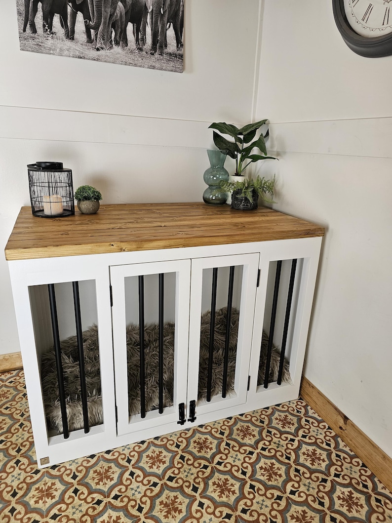 K&ouml;nnte beinhalten: Eine wei&szlig;e Hundebox mit einer Holzplatte und schwarzen St&auml;ben. Die Box hat vier T&uuml;ren und ein flauschiges Bett im Inneren. Die Box steht in einer Ecke mit gemustertem Boden und dekorativen Gegenst&auml;nden.