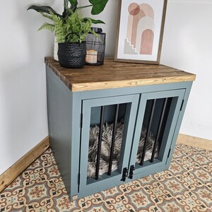 K&ouml;nnte beinhalten: Eine blaue Hundebox mit einer Holzplatte, mit schwarzen St&auml;ben an den Vordert&uuml;ren. Die Box hat einen flauschigen Innenraum und ist mit einem gerahmten abstrakten Kunstdruck, einem schwarzen geflochtenen Korb mit Pflanzen und einer schwarzen Laterne versehen.