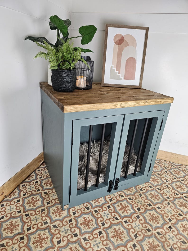 K&ouml;nnte beinhalten: Eine blaue Hundebox mit einer Holzplatte, mit schwarzen St&auml;ben an den Vordert&uuml;ren. Die Box hat einen flauschigen Innenraum und ist mit einem gerahmten abstrakten Kunstdruck, einem schwarzen geflochtenen Korb mit Pflanzen und einer schwarzen Laterne versehen.