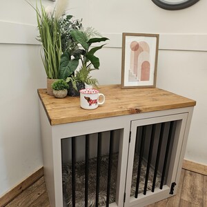 K&ouml;nnte beinhalten: Ein Hundezwinger aus Holz mit grauem Rahmen und zwei vergitterten T&uuml;ren. Die Oberseite ist aus Holz und der Innenraum hat ein flauschiges Bett. Eine Tasse mit einem Hunde-Design, Pflanzen und ein gerahmtes Bild stehen oben. Im Hintergrund ist eine Uhr zu sehen.