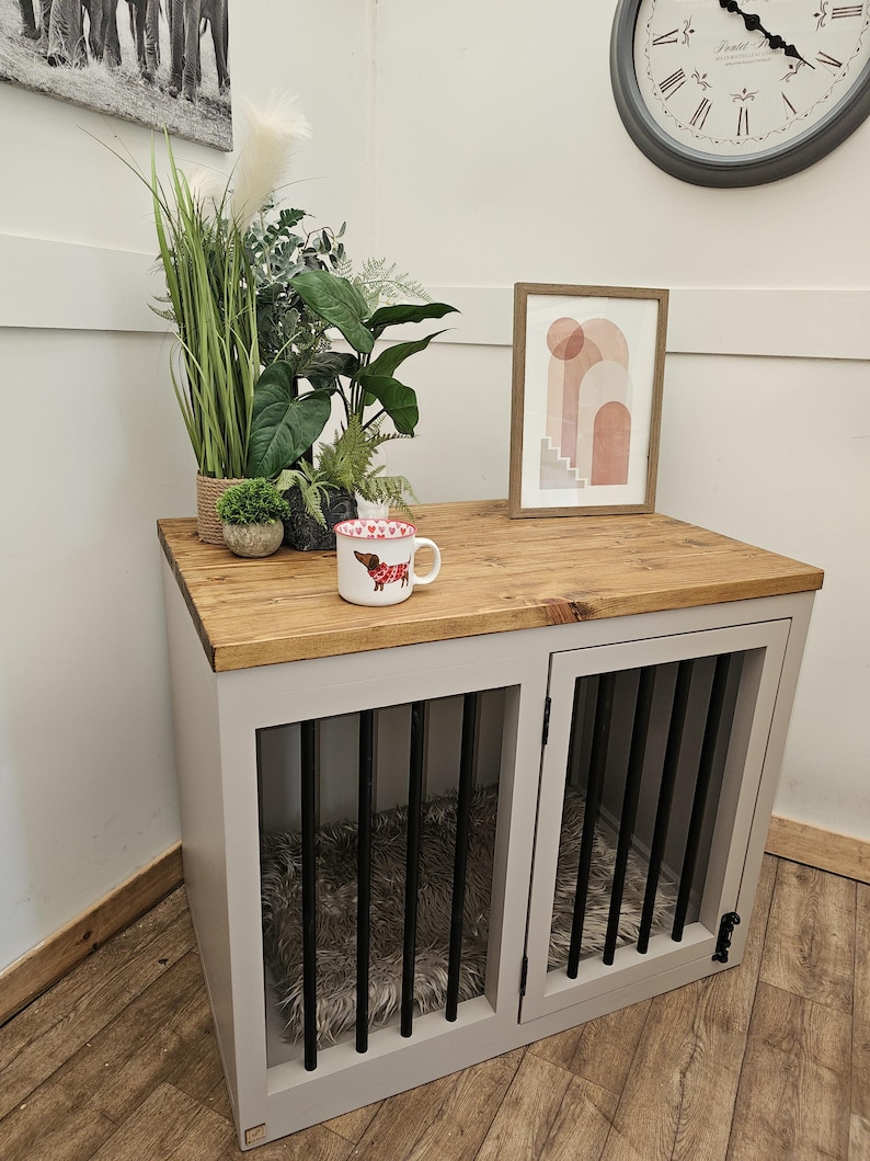 K&ouml;nnte beinhalten: Ein Hundezwinger aus Holz mit grauem Rahmen und zwei vergitterten T&uuml;ren. Die Oberseite ist aus Holz und der Innenraum hat ein flauschiges Bett. Eine Tasse mit einem Hunde-Design, Pflanzen und ein gerahmtes Bild stehen oben. Im Hintergrund ist eine Uhr zu sehen.