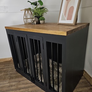 K&ouml;nnte beinhalten: Eine schwarze Hundebox mit einer Holzplatte und Metallst&auml;ben. Die Box hat drei T&uuml;ren und ein flauschiges Bett im Inneren. Dekorative Gegenst&auml;nde, darunter Pflanzen und ein gerahmter Druck, befinden sich oben auf der Box.
