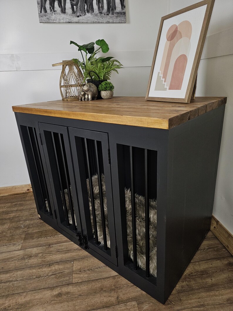K&ouml;nnte beinhalten: Eine schwarze Hundebox mit einer Holzplatte und Metallst&auml;ben. Die Box hat drei T&uuml;ren und ein flauschiges Bett im Inneren. Dekorative Gegenst&auml;nde, darunter Pflanzen und ein gerahmter Druck, befinden sich oben auf der Box.