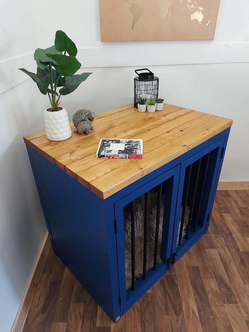 K&ouml;nnte beinhalten: Eine blaue Hundebox mit einer Oberseite aus Naturholz. Die Box hat zwei T&uuml;ren mit schwarzen St&auml;ben. Eine wei&szlig;e Vase mit einer gr&uuml;nen Pflanze, eine Igel-Figur und ein kleines Buch befinden sich oben. Eine schwarze Laterne und kleine Topfpflanzen befinden sich ebenfalls oben.