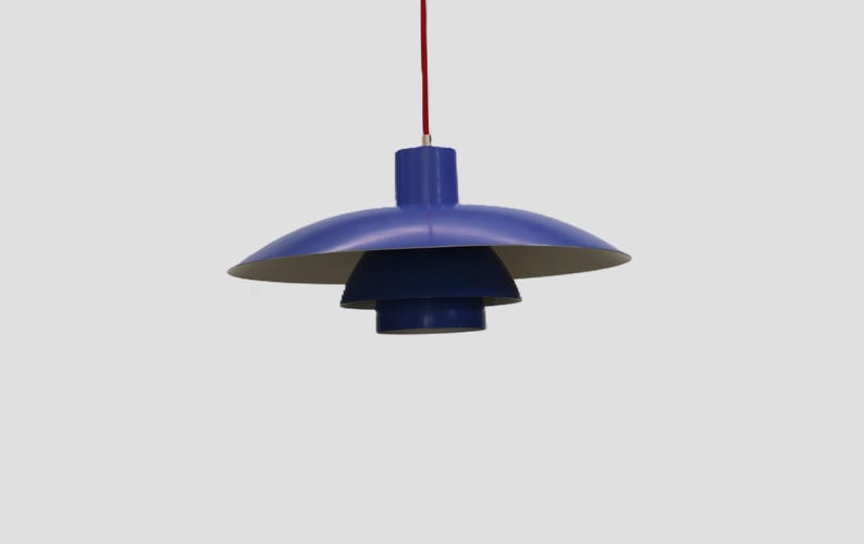 Puede incluir: Una moderna l&aacute;mpara colgante azul con un dise&ntilde;o en capas. La l&aacute;mpara tiene una pantalla redondeada y una secci&oacute;n inferior escalonada, suspendida por un cable rojo. El interior de la pantalla es de color claro.