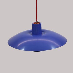 Puede incluir: Una l&aacute;mpara colgante azul con una pantalla redondeada. La l&aacute;mpara cuelga de un cable rojo. La l&aacute;mpara tiene una parte superior cil&iacute;ndrica y una pantalla ancha, poco profunda, en forma de cuenco. Fondo neutro.