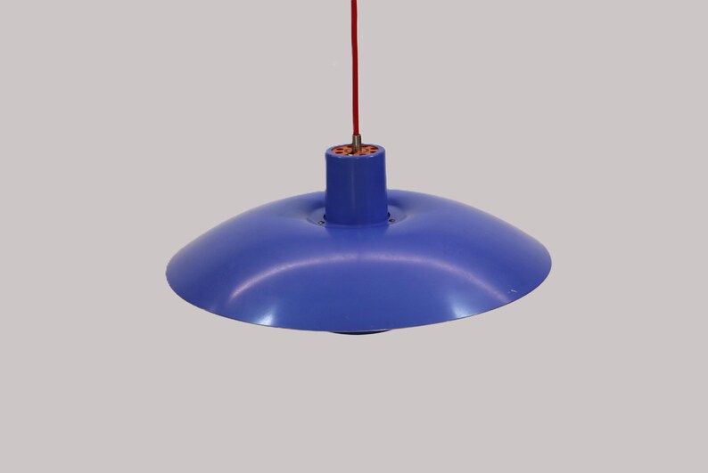 Puede incluir: Una l&aacute;mpara colgante azul con una pantalla redondeada. La l&aacute;mpara cuelga de un cable rojo. La l&aacute;mpara tiene una parte superior cil&iacute;ndrica y una pantalla ancha, poco profunda, en forma de cuenco. Fondo neutro.