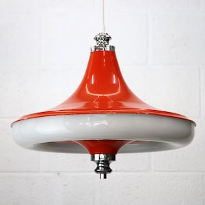 Puede incluir: Lámpara colgante de estilo retro con una parte superior naranja brillante y una base circular blanca y ancha. El accesorio tiene un elemento decorativo cromado en la parte superior e inferior. Cuelga de un cable blanco.