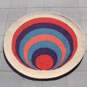 May include: A round rug with a concentric circle design. The circles are in shades of red, orange, blue, and purple.