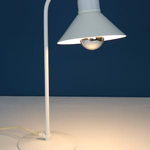 Può includere: Lampada da tavolo bianca con paralume bianco e base bianca. La lampada ha un braccio curvo e una lampadina.