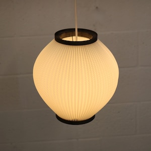 May include: A cream-coloured, pleated lampshade in a rounded shape, suspended from a white cord. The shade is framed by black rings at the top and bottom. The background is a white brick wall.