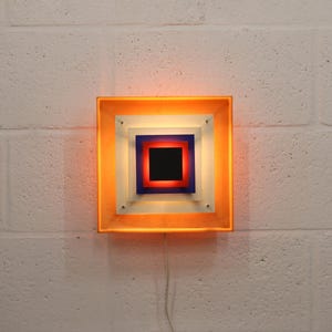 May include: A square, retro-style wall sconce with a layered design. The outer frame is orange, surrounding white, blue, and red squares. The center is black. The light fixture is mounted on a white brick wall.