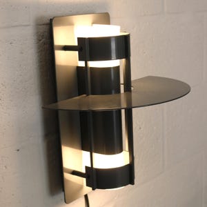 May include: A modern, black wall sconce with a built-in shelf. The sconce features a cylindrical design with horizontal bands of light. The fixture is mounted on a rectangular backplate and has a curved shelf. A black power cord extends from the bottom.