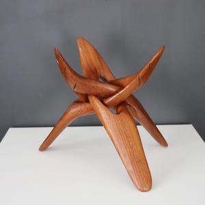 Puede incluir: Un soporte de trípode de madera con un diseño único y abstracto. El soporte está hecho de tres piezas de madera que se entrelazan para crear una base estable.